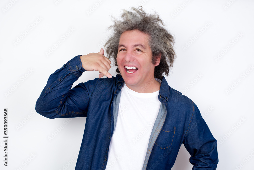 Handsome middle aged Caucasian man with afro gray hair, against white wall, imitates telephone conversation, makes phone call gesture with hands, has confident expression. Call me! Handsome middle aged Caucasian man with afro gray hair, against white wall, imitates telephone conversation, makes phone call gesture with hands, has confident expression. Call me!