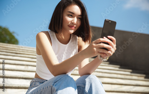 Fotografie Happy asian woman sitting on street stairs and taking selfie, smiling at mobile phone camera