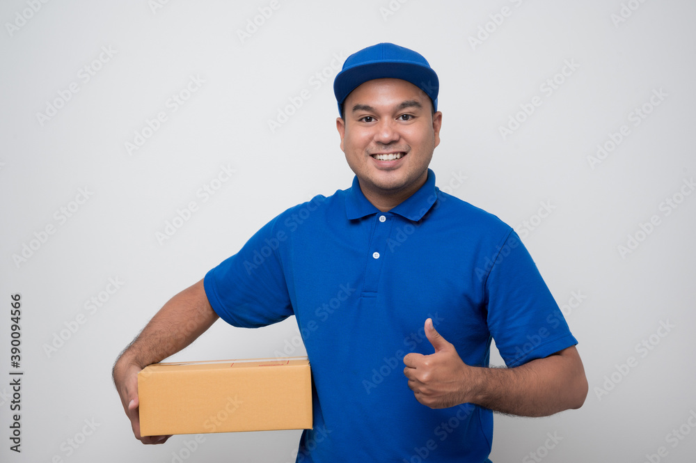 Young smiling asian delivery man in blue uniform holding box parcel cardboard and showing thumbs up on isolated white background. Young smiling asian delivery man in blue uniform holding box parcel cardboard and showing thumbs up on isolated white background.