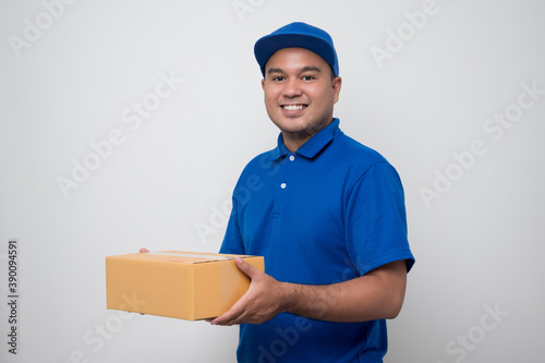 Young smiling asian delivery man in blue uniform holding box parcel cardboard on isolated white background.