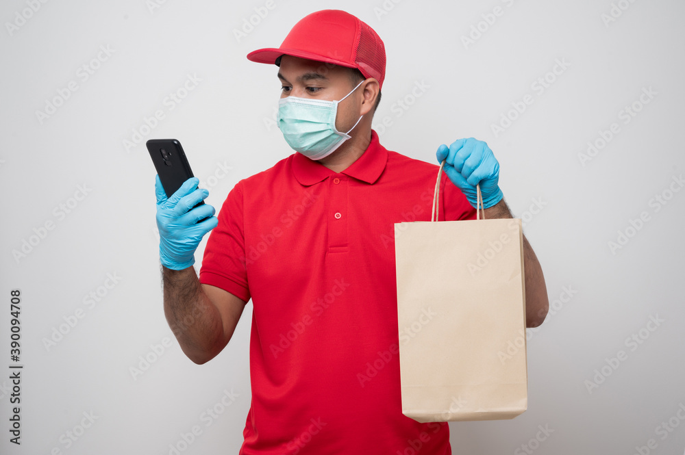 Young asian delivery man in red uniform wearing protection mask and medical rubber gloves using smartphone connecting with customer to delivery parcel paper bag or food delivery.