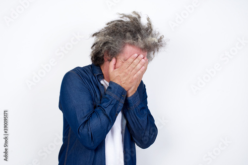 Sad Handsome middle aged Caucasian man with afro gray hair, against white wall,  covering face with hands and crying.