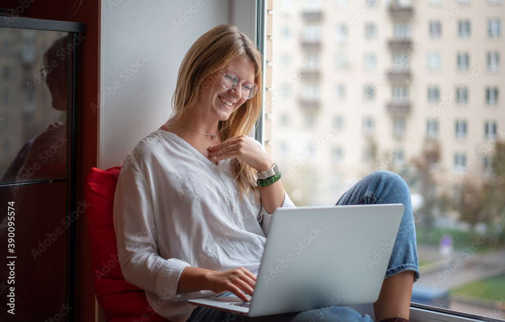 Young woman with long red hair wearing glasses in white shirt works on a laptop at home, freelancer and remote business. Sitting on the windowsill Young woman with long red hair wearing glasses in white shirt works on a laptop at home, freelancer and remote business. Sitting on the windowsill