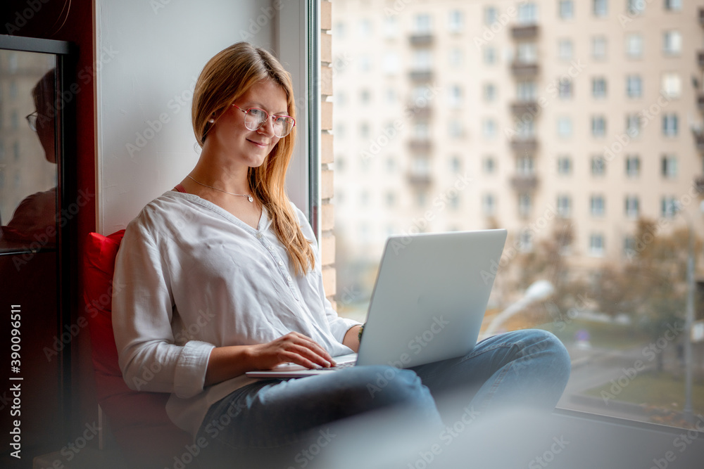 Young woman with long red hair wearing glasses in a white shirt works on a laptop at home, freelancer and remote business. Sitting on the windowsill Young woman with long red hair wearing glasses in a white shirt works on a laptop at home, freelancer and remote business. Sitting on the windowsill