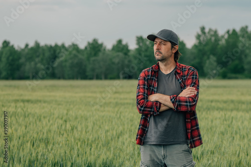 Wheat farmer agronomist standing in cultivated cereal crops agricultural field with arms crossed and looking into distance with uncertain facial expression