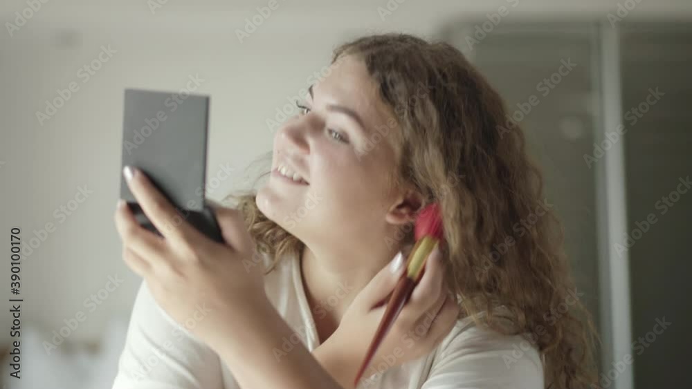 Close-up of confident young plus-size woman applying make-up and smiling. Portrait of happy plump Caucasian lady with face powder and hand mirror smiling. Beauty and confidence concept. Close-up of confident young plus-size woman applying make-up and smiling. Portrait of happy plump Caucasian lady with face powder and hand mirror smiling. Beauty and confidence concept.