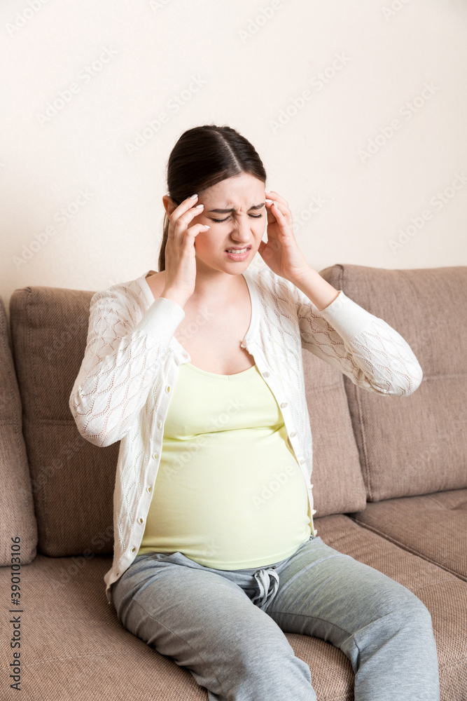 Young pregnant woman with headache sitting on sofa. Pregnancy expectation concept Young pregnant woman with headache sitting on sofa. Pregnancy expectation concept