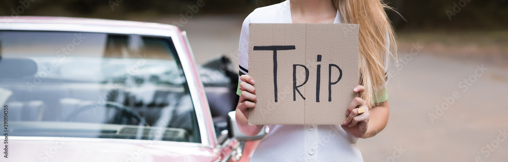 cropped view of woman holding card with trip lettering near vintage cabriolet on blurred background, banner