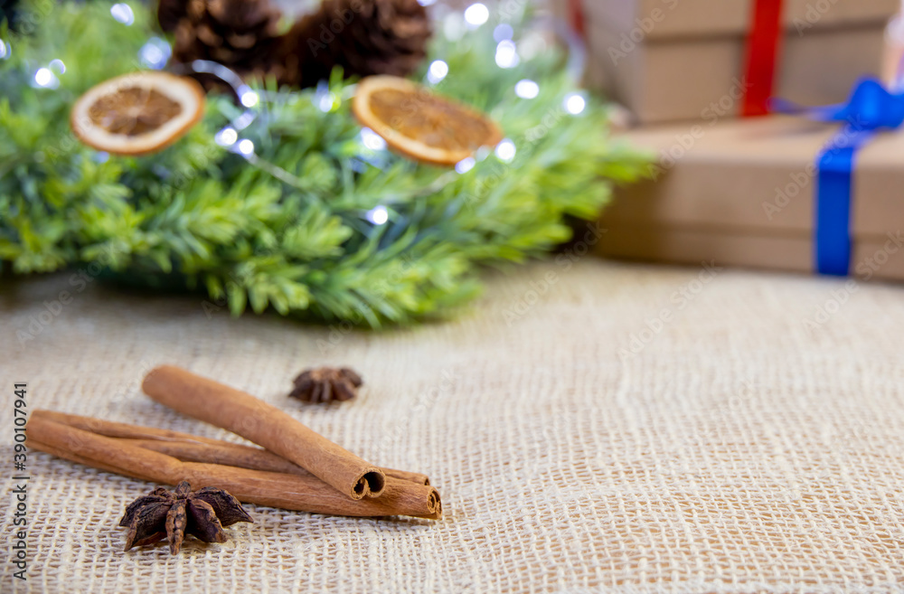 
close-up of cinnamon stick, anise, green wreath with lights, dry oranges and cone, two gifts with colorful ribbons and bows, bright christmas decoration on jute background, cardboard box, front view