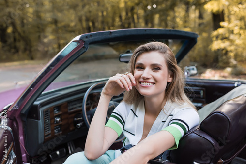 happy young woman looking at camera while sitting in vintage cabriolet