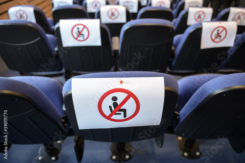 Shallow depth of field with social distance warning signs on seats during a show, concert or play in a theater or cinema, conference hall. Empty rows of places marked with a warning 'don't sit' sign.