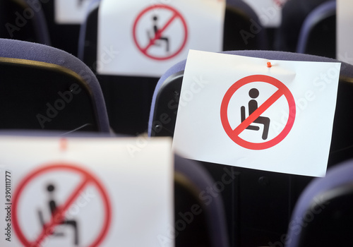 Shallow depth of field with social distance warning signs on seats during a show, concert or play in a theater or cinema, conference hall. Empty rows of places marked with a warning 'don't sit' sign.