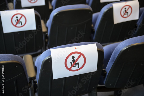 Shallow depth of field with social distance warning signs on seats during a show, concert or play in a theater or cinema, conference hall. Empty rows of places marked with a warning 'don't sit' sign.