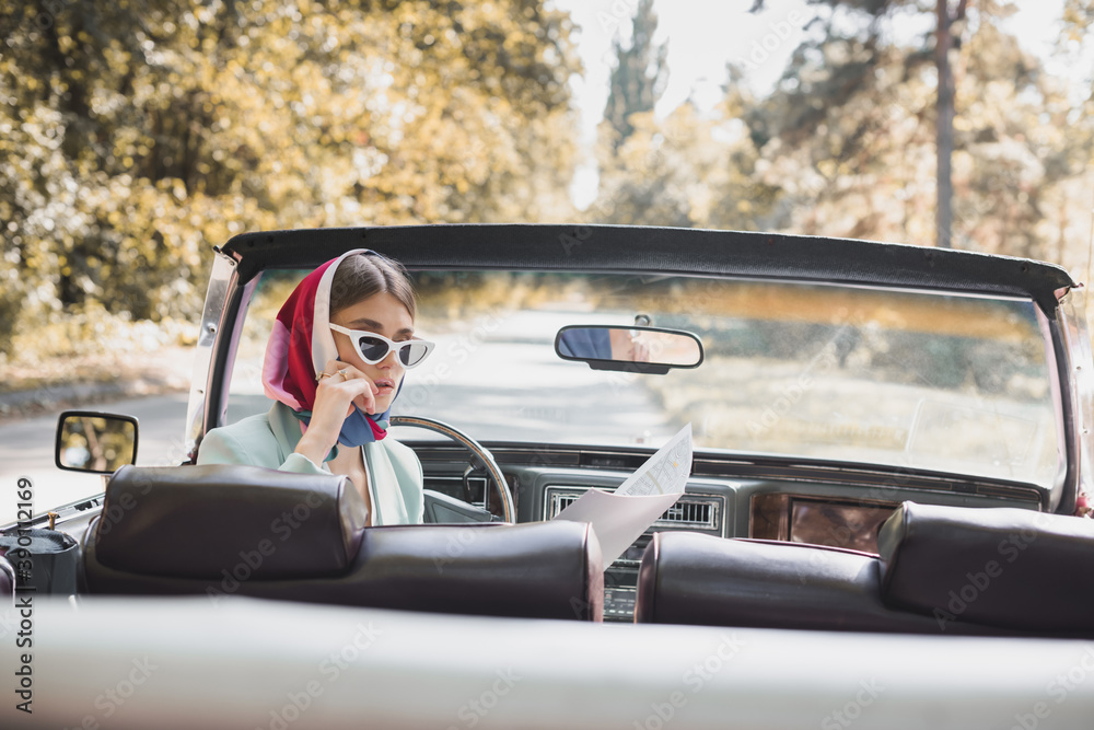 Pensive woman in sunglasses looking at map on driver seat of vintage car on blurred foreground