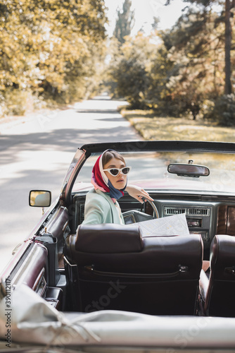 Stylish woman with map sitting in auto on blurred foreground on road during trip