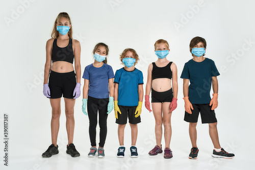 Full length shot of five sportive kids in sportswear wearing medical masks and protective gloves looking at camera, standing together isolated over white background