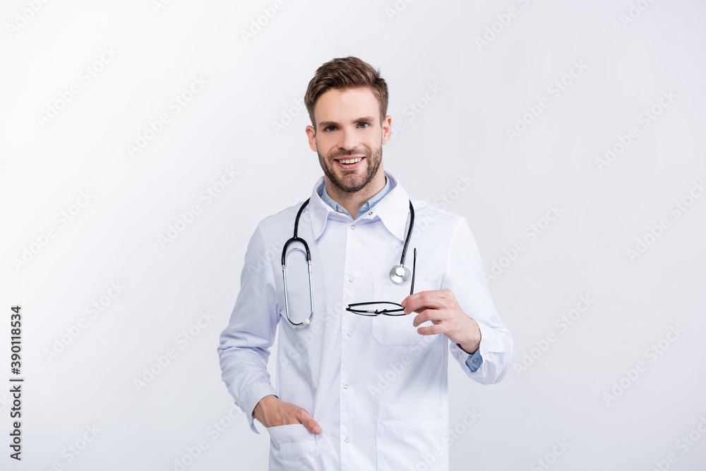 Front view of smiling ophthalmologist with hand in pocket holding eyeglasses, while looking at camera isolated on white Front view of smiling ophthalmologist with hand in pocket holding eyeglasses, while looking at camera isolated on white