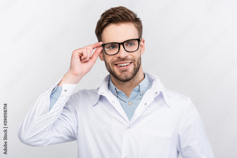 Smiling doctor in white coat touching eyeglasses isolated on grey Smiling doctor in white coat touching eyeglasses isolated on grey