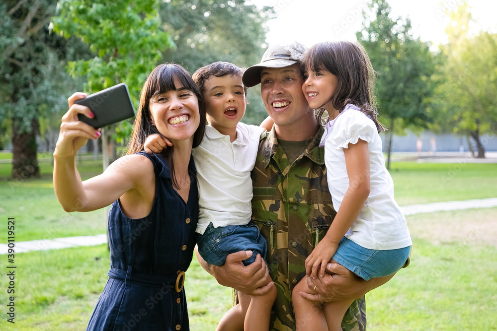 Fototapeta premium Happy excited military man, his wife and two kids celebrating dads returning, enjoying leisure time in park, taking selfie on cell phone. Medium shot. Family reunion or returning home concept