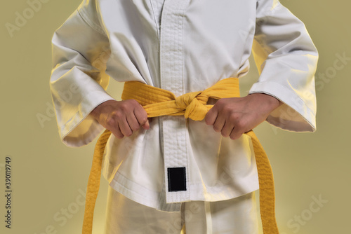 Cropped shot of little karate girl in white kimono adjusting yellow belt while getting ready for exercising, doing martial arts, standing isolated over yellow background