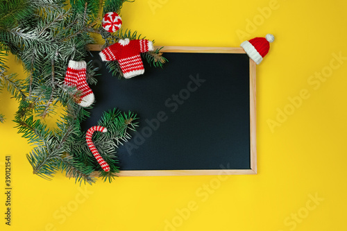 black chalk board, fir-tree branch and decorative knitted ornaments sock, sweater, hat and caramel in red and white tone on yellow background photo