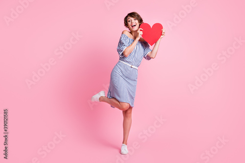 Photo portrait of lovely girl standing on one leg holding big red heart postcard isolated on pastel pink colored background © deagreez