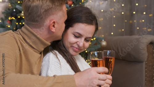 Young couple in love sitting and hugging near the Christmas tree, drinking a glass of champagne, while he whispers to her how much he loves her.
