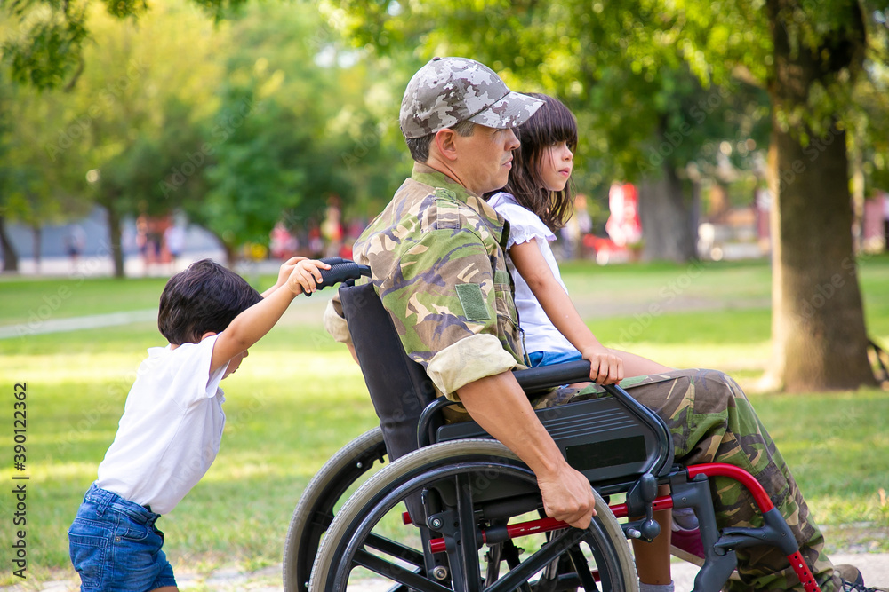 Disabled military veteran walking with two children in park. Girl sitting on dads lap, boy pushing wheelchair. Veteran of war or disability concept