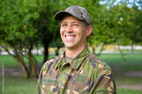 Portrait of happy man in military camouflage uniform standing in park, looking at camera and smiling. Green trees in background. Military man or guard concept
