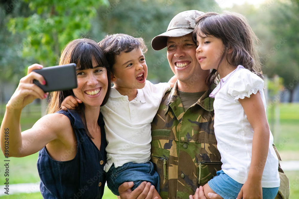 Fototapeta premium Happy joyful military family celebrating dads returning, enjoying leisure time in park, taking selfie on smartphone. Medium shot. Family reunion or returning home concept