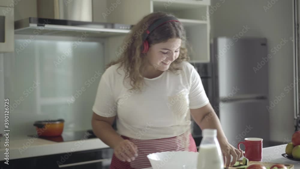 Cheerful plump woman in headphones dancing in kitchen and eating fresh healthful cucumber. Portrait of joyful young Caucasian obese lady enjoying sunny weekend morning at home.