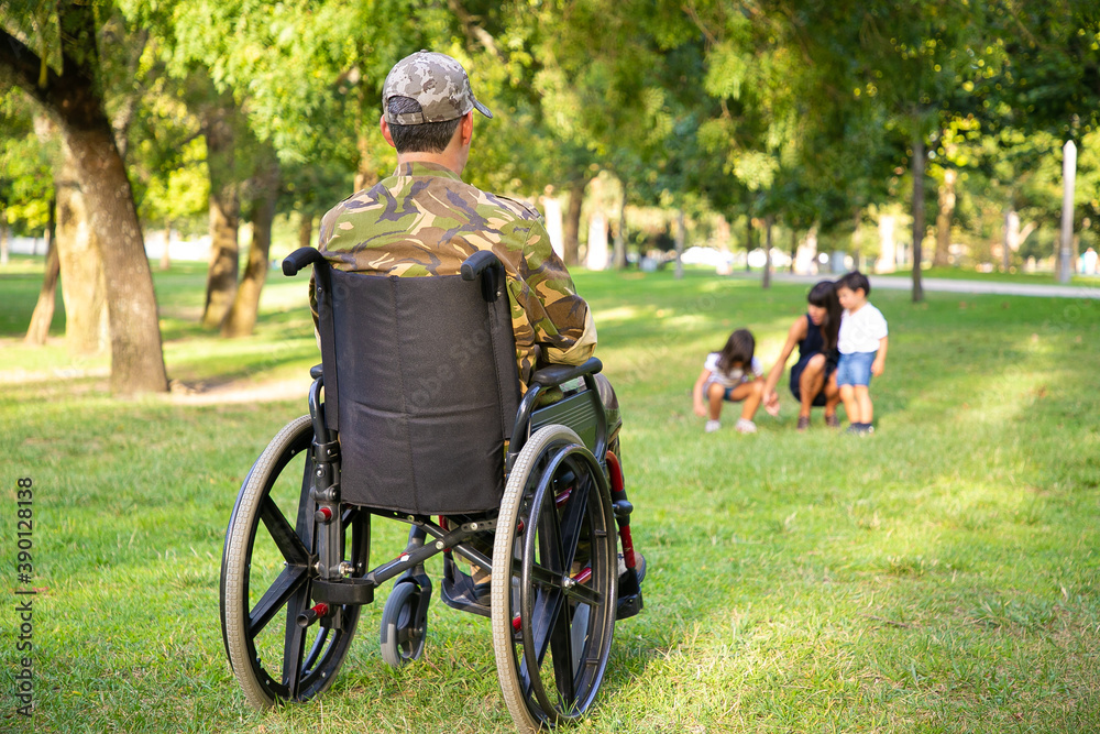 Lonely disabled retired military man in wheelchair looking at his wife and little children playing together in park. Back view. Veteran of war or disability concept Lonely disabled retired military man in wheelchair looking at his wife and little children playing together in park. Back view. Veteran of war or disability concept