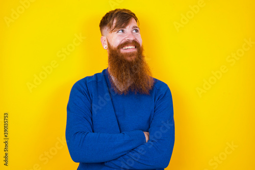 Young handsome red haired bearded man standing against yellow wall, with thoughtful expression, looks away keeps hands down bitting his lip thinks about something pleasant.