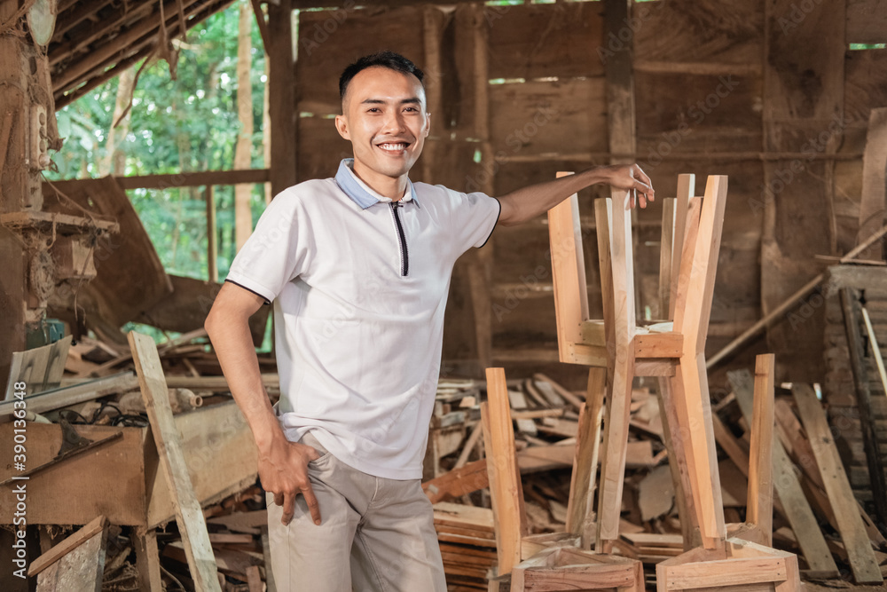 The young entrepreneur smiles with hand resting on wooden chairs while standing at the workshop