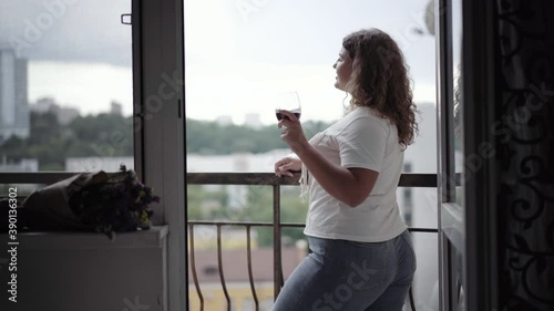 Side view of happy young chubby woman enjoying overcast evening on balcony. Portrait of relaxed Caucasian lady drinking wine from glass and smiling. Positivity and lifestyle. photo