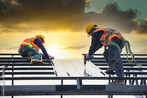 Asian construction workers wear safety straps while working on the roof structure of the building at a construction site. Roofer, using a pneumatic nail gun, install roof tiles.