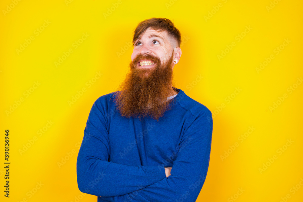 Portrait of mysterious Young handsome red haired bearded man standing against yellow wall looking up with enigmatic smile. Advertisement concept.