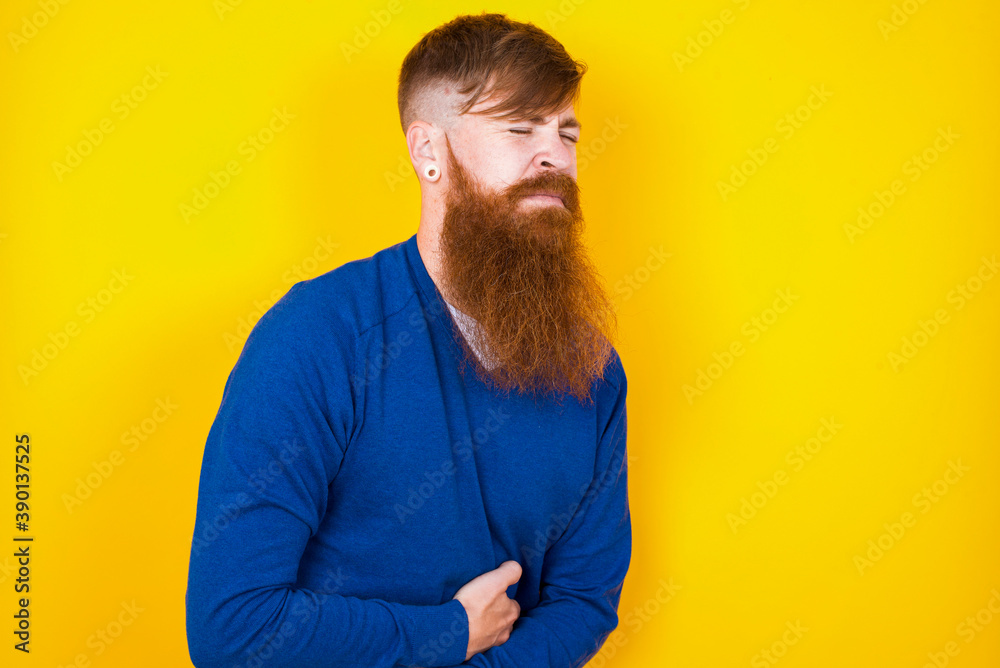 Young handsome red haired bearded man standing against yellow wall suffering from strong stomachache. Young handsome red haired bearded man standing against yellow wall suffering from strong stomachache.