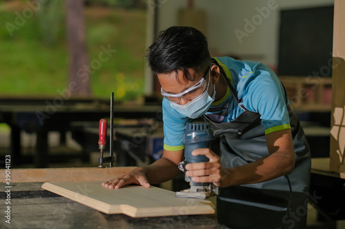Young carpenter in work clothes and face mask using trimmer for console table