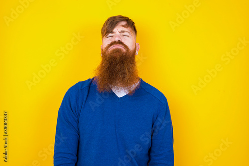 Gloomy, bored Young handsome red haired bearded man standing against yellow wall frowns face looking up, being upset with so much talking hands down, feels tired and wants to leave.
