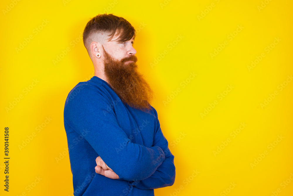 Image of upset Young handsome red haired bearded man standing against yellow wall with arms crossed. Looking with disappointed expression aside after listening to bad news. Image of upset Young handsome red haired bearded man standing against yellow wall with arms crossed. Looking with disappointed expression aside after listening to bad news.