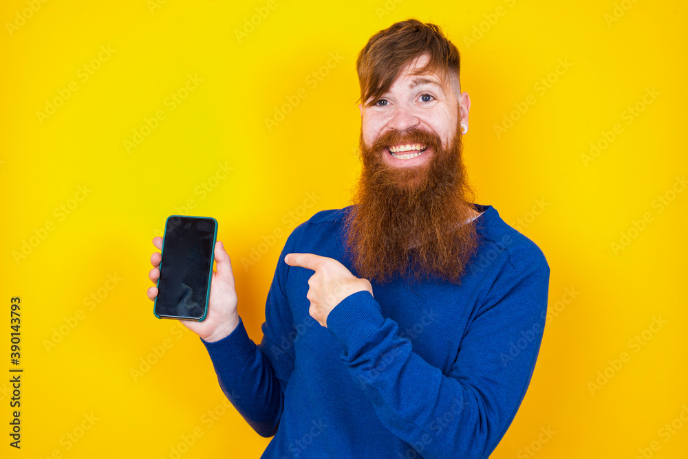 Excited Young handsome red haired bearded man standing against yellow wall holding and pointing with finger at smartphone with blank screen. Advertisement concept. Excited Young handsome red haired bearded man standing against yellow wall holding and pointing with finger at smartphone with blank screen. Advertisement concept.