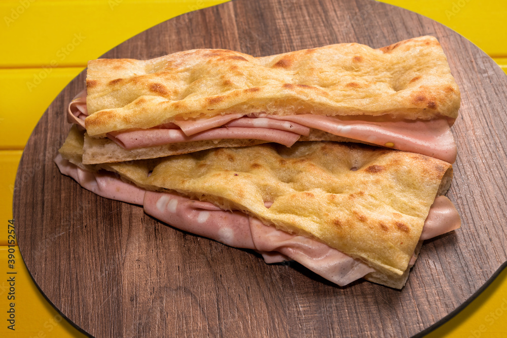 two focaccia bread sandwich filled with bologna mortadella on wooden cutting board and yellow background