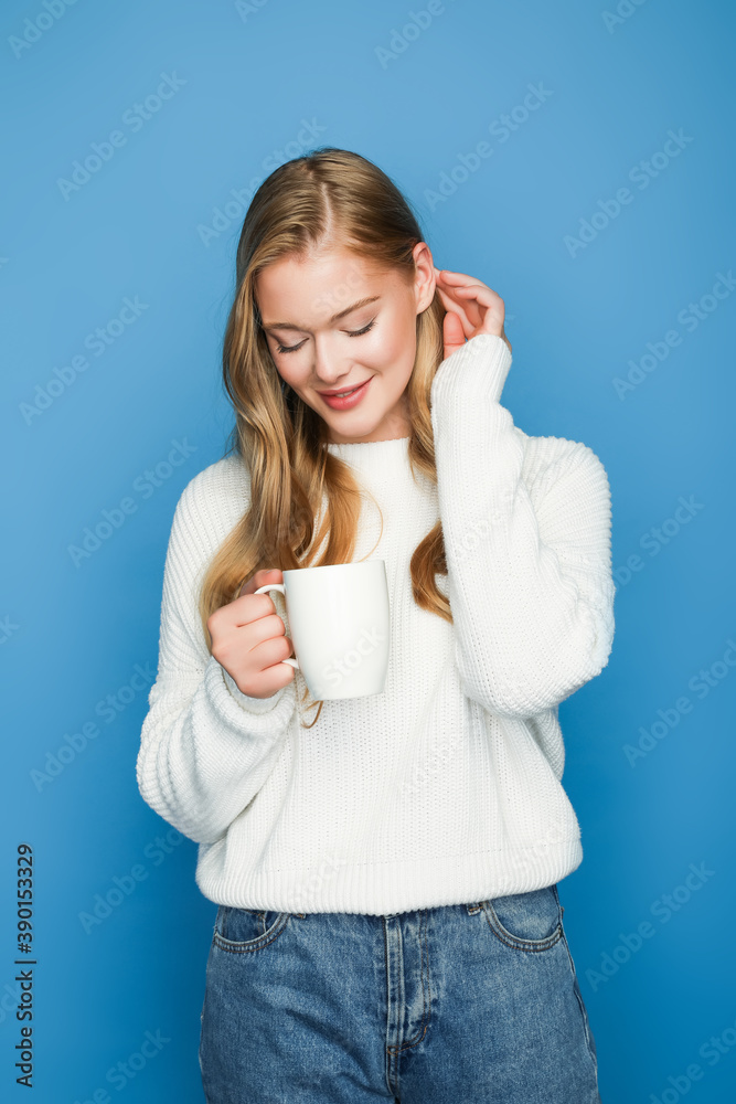 happy blonde beautiful woman in sweater pointing at mug isolated on blue background happy blonde beautiful woman in sweater pointing at mug isolated on blue background