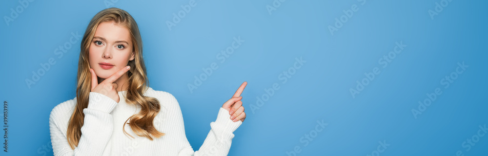pensive blonde beautiful woman in sweater pointing with finger isolated on blue, banner pensive blonde beautiful woman in sweater pointing with finger isolated on blue, banner
