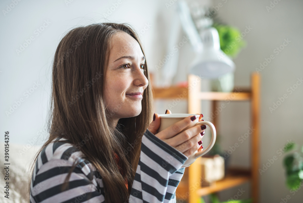 A young beautiful girl in pajamas is sitting on the bed on a sunny morning and enjoying a cup of aromatic coffee. Concept of a nice start to the day A young beautiful girl in pajamas is sitting on the bed on a sunny morning and enjoying a cup of aromatic coffee. Concept of a nice start to the day