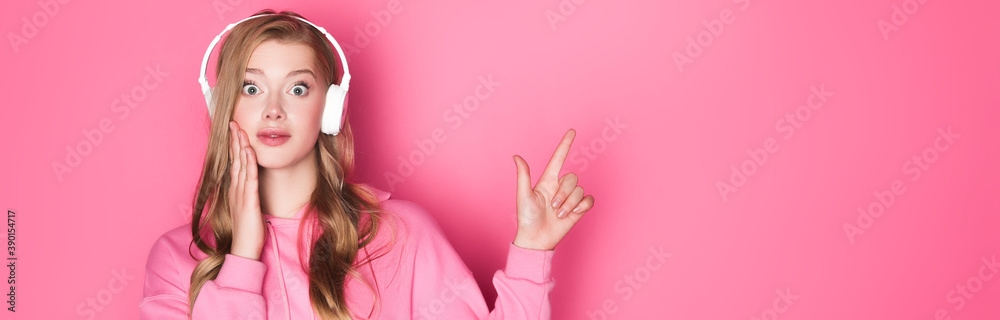 shocked beautiful woman listening music in headphones and pointing up on pink background, banner - obrazy, fototapety, plakaty 