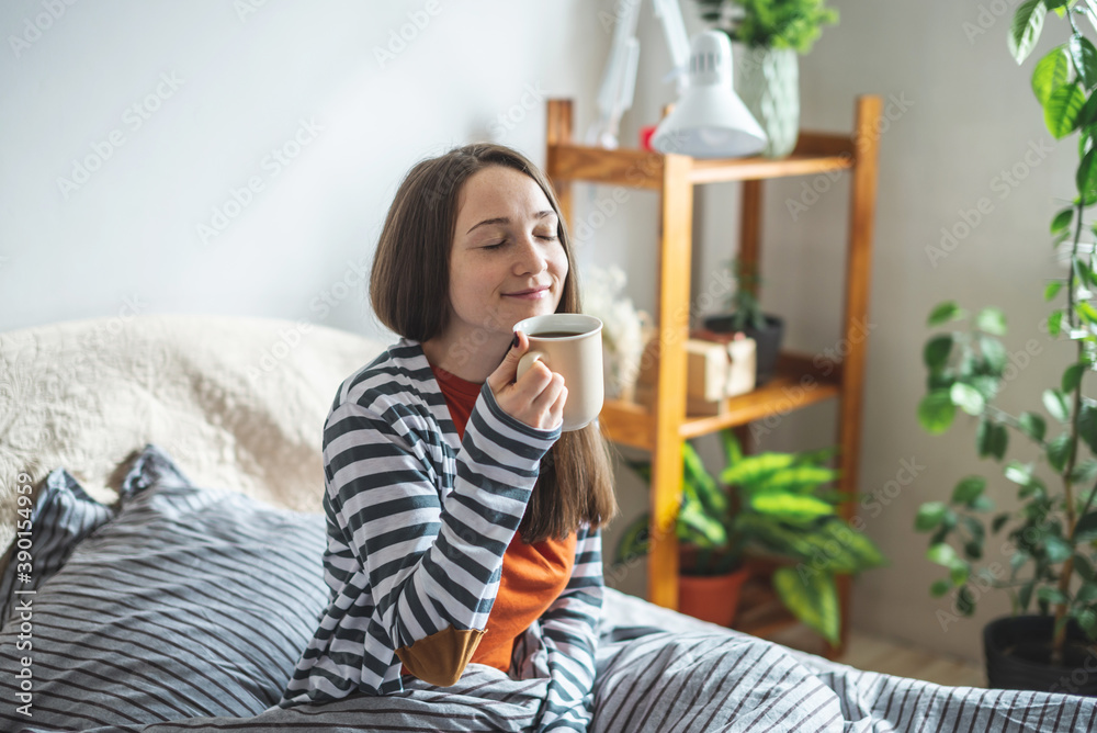 A young beautiful girl in pajamas is sitting on the bed on a sunny morning and enjoying a cup of aromatic coffee. Concept of a nice start to the day A young beautiful girl in pajamas is sitting on the bed on a sunny morning and enjoying a cup of aromatic coffee. Concept of a nice start to the day