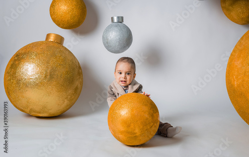a beautiful little boy in a suit with a bow tie sits with a Golden ball on a white background with giant holiday balloons