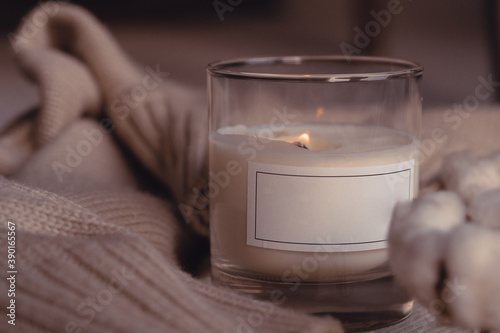 Symbol Of Coziness. Burnng Scented Aroma Candle In Glass With Blank White Label Near Cotton Flower On Cozy Stylish Sweater photo